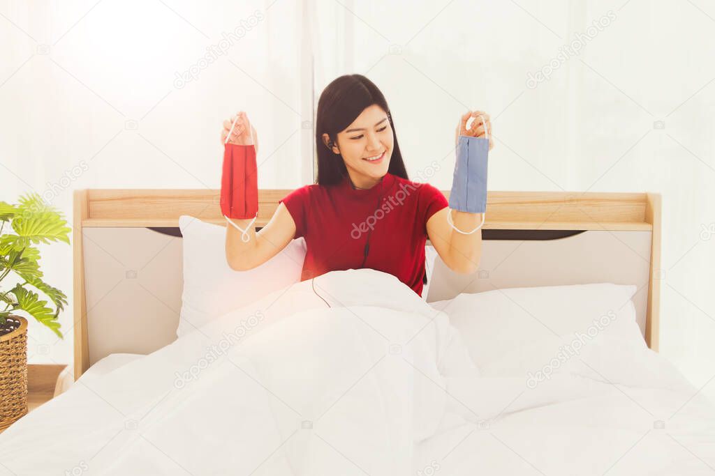 Normal new concepts:Portrait of a beautiful Asian woman wearing a red shirt, smiling comfortably on her bed at home, carrying a red and blue cloth mask suitable for traveling to work.