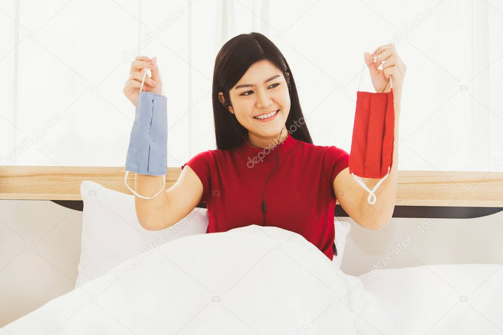 Concept new normal : Portrait young beautiful Asian woman wearing a red shirt, smiling on the bed, choosing a new red and blue mask that is pleasing, clean for work.