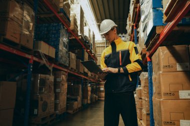 Güvenlik kaskı takan profesyonel bir erkek işçi endüstriyel depolama deposunda çalışıyor ve elektronik ekipman taşıma lojistiği kullanarak depolama ve dağıtım malzemelerini depoluyor..
