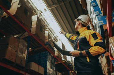 Güvenlik kaskı takan profesyonel bir erkek işçi endüstriyel depolama deposunda çalışıyor ve elektronik ekipman taşıma lojistiği kullanarak depolama ve dağıtım malzemelerini depoluyor..