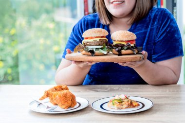 Asyalı şişman kadınlar, hazırladığı kızarmış tavukla, hamburgerle ve pizzayla yemek yemekten memnuniyet duyarlar bu resim hamburger, kolesterol ve aşırı yağ kavramına odaklanır..