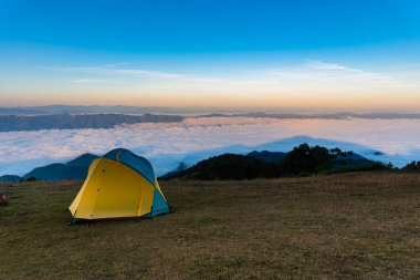 Sarı kamp çadırı yüksek bir dağda, dağ kompleksi manzaralı ve yoğun beyaz sis sabah güneş ışığına yansıyan, turist ve seyahat konseptine.
