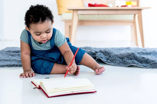 A 1 year old half-African half-Thai girl, Sitting on the floor And using a pencil to write on the notebook, to child development and education concept.