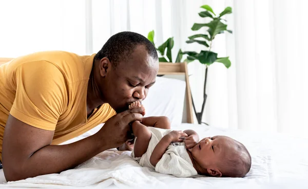 An African American father kissing foot, his 12-day-old baby newborn, is laying on the white bed, with love and happy, concept to African American family and baby black skin newborn