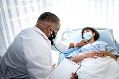 Afrikalı Amerikalı doktor cerrahi maske takıyor. Steteskop kullanıyor. Asyalı kadın hastaların semptomlarını, sağlık hizmetlerini ve sağlık sigortası kavramlarını dinleyip kontrol ediyorlar.