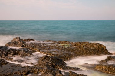 Güzel kayalık plaj, sabah güneşinin ilk ışıklarıyla ışıldıyor. Altından güneş ışığı pürüzsüz deniz suyuna yansıyor.) 