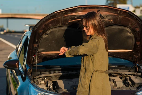 Young lady standing near her broken car on the highway roadside, checking the time and getting stressed about being late — Stockfoto