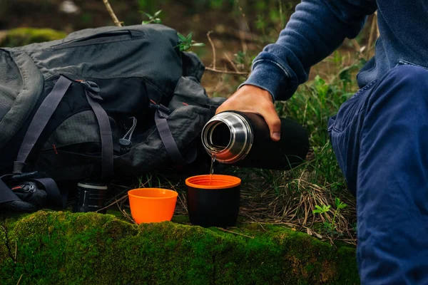 Drinking tea during hike. Man hand pouring hot tea from black thermos bottle into 2 cups in the woods. Refreshment during hiking. Camping equipment — Stock Photo, Image