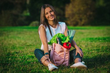 Güzel vejetaryen kadın çimlerin üzerinde pembe, yeniden kullanılabilir, biyolojik sebzeli bir alışveriş çantasıyla oturuyor. Bitki temelli vegan diyeti. Sağlıklı yiyecekler temiz yiyecekler. Sıfır atık, plastiksiz konsept