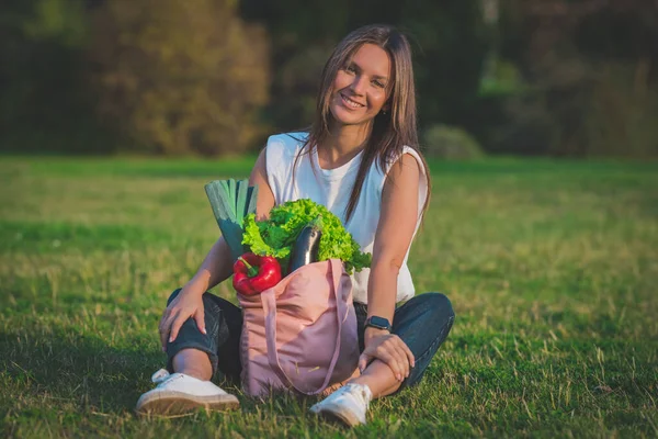 Hermosa mujer joven vegana sentada en la hierba con rosa reutilizable bolsa de compras textil con verduras bio frescas. Dieta vegana a base de plantas. Comida sana, comida limpia. Cero residuos, concepto libre de plástico — Foto de Stock