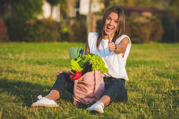 Hermosa mujer vegana sonriente feliz sentado en la hierba con rosa eco reutilizable bolsa de comestibles con verduras bio frescas. Dieta a base de plantas. Comida sana, comida limpia. Cero residuos, concepto libre de plástico — Foto de Stock