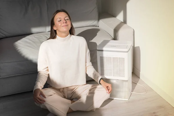 Woman breathing fresh air at home. Air purifier in a living room. Dehumidifier, humidity indicator, air ionizer or water container. — Stock Photo, Image