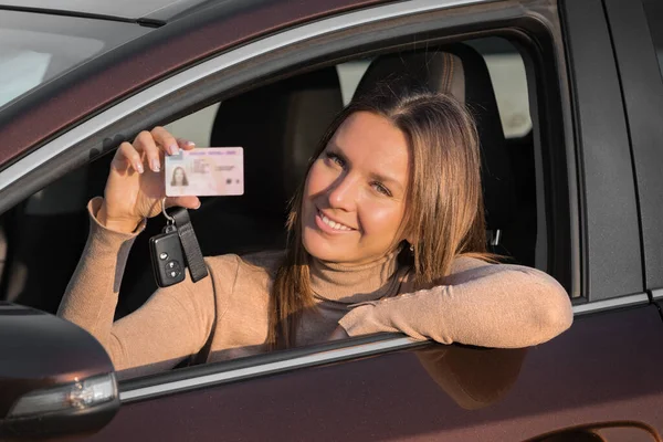 迷人的微笑的年轻女子骄傲地从车窗向外展示她的驾驶执照。女人已经拿到了驾驶执照，感到非常高兴和兴奋。准备开车 — 图库照片