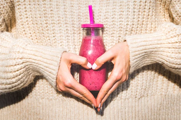 Frau im Wollpullover, die mit ihren Händen Herzform herstellt und eine Flasche mit rosa selbstgemachtem Detox-Blueberry-Himbeer-Smoothie oder -Saft in der Hand hält. Gesunde, saubere Ernährung — Stockfoto
