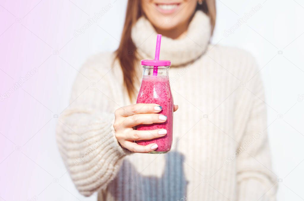Smiling woman in woollen sweater holding bottle with pink homemade detox blueberry and raspberry smoothie or juice. Healthy clean eating