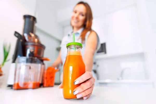 Happy healthy woman making fresh carrot juice in a juicer in the kitchen at home. Bottle of fresh homemade carrot juice. Healthy lifestyle. Clean eating, detox concept — Stok Foto