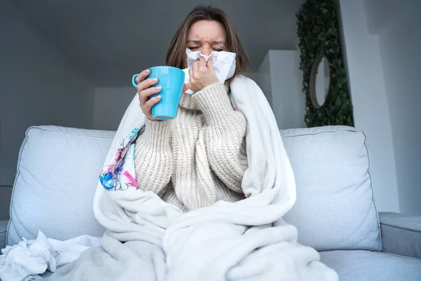 Jovem doente com xícara de bebida quente sentado no sofá coberto com cobertor em casa. Nariz a soprar com guardanapo. Vírus da gripe, gripe, constipação. — Fotografia de Stock