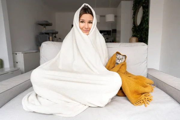 Una mujer con frío. La mujer que se congela y el gato bajo la tela a cuadros tienen frío en el sofá en casa. Calentamiento bajo una manta en frío otoño o invierno. —  Fotos de Stock