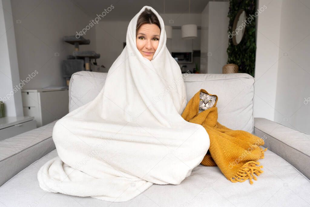Woman feeling cold. Freezing woman and cat under a plaid have cold on the sofa at home. Warming under a blanket in cold autumn or winter.