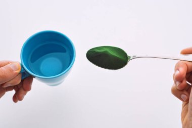 Klorella, spirulina ya da buğday çimi içeren bir bardak su ve kaşık tutan erkek eli. Detoks sıvısı hazırlığı. Yeşil gıda takviyesi, klorofil, süper yiyecek.