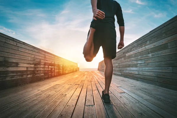 젊고 날씬 한 남자가 태어 나거나 일출 때 밖으로 뛰기 전에 몸을 따뜻하게 하고 다리를 쭉 뻗는다. 검은 운동복을 입은 운동 선수가 건강 운동을 하고 있습니다. 스포츠와 건강 한 생활 방식. — 스톡 사진