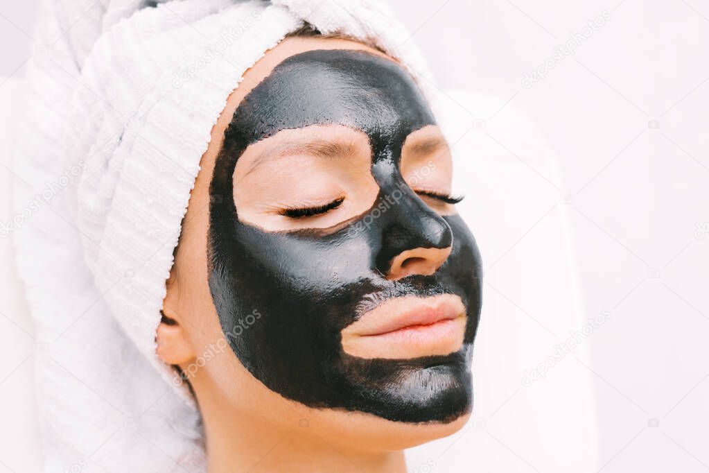 Portrait of beautiful woman with towel on her head relaxing with black purifying charcoal facial mask in spa. Cosmetology and skincare. Beauty treatments. Spa therapy.