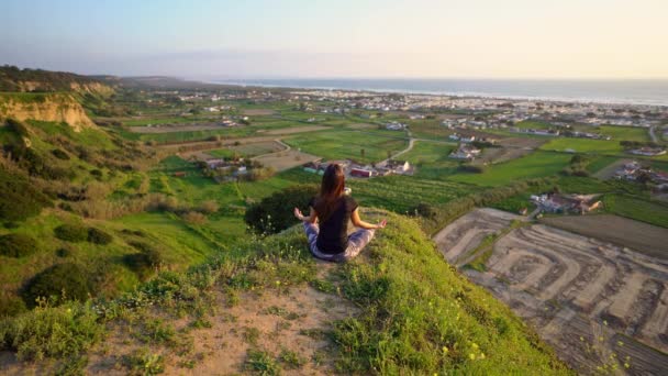 Kvinna utövar yoga och mediterar på toppen av berget med vacker utsikt över staden och havet vid solnedgången. Kvinna som sitter i lätt pose eller sukhasana med mudra. Avkoppling, harmoni med naturen — Stockvideo