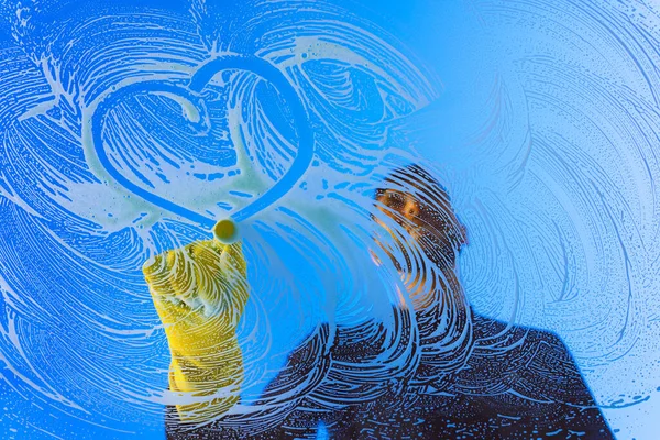Limpiador de ventanas o lavadora con guantes amarillos dibujando la forma del corazón en un cristal de ventana. Corazón de amor símbolo dibujado en vidrio. Concepto de limpieza de ventanas — Foto de Stock
