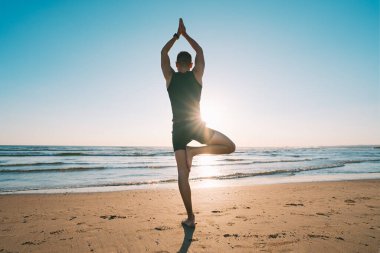 Sahilde gün batımında ya da gün doğumunda okyanus manzaralı yoga yapan bir adam. Ağaç duruşu ya da elleri yukarıda vrksasana. Rahatlama, doğayla uyum. Sağlıklı yaşam tarzı