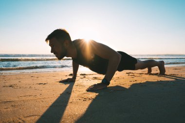 Young fit man doing push ups or exercising on the beach during sunrise in the morning. Athletic man doing fitness workout or training outdoors. Sports and healthy lifestyle