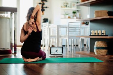Evde elmas pozisyonunda oturan, kollarını açıp internetteki özel derslere bakan bir kadın..