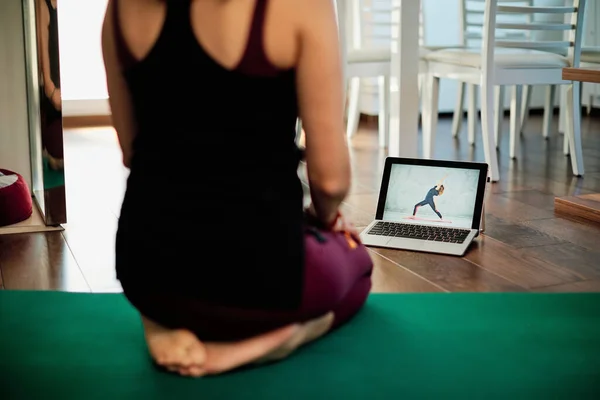 Rear view of woman sitting on yoga mat at home and looking at yoga tutorials on internet.