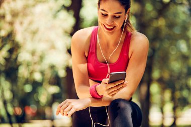 Gülümseyen kadın koşucu doğada mola veriyor ve bir telefon kullanıyor..