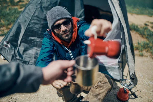 Young bearded man crouching next to the tent on a camping trip and pouring coffee into the mug.