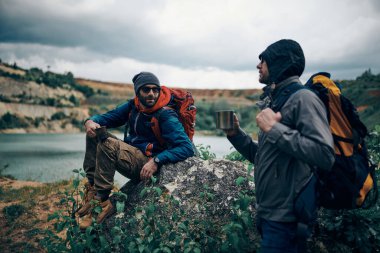 Doğada bir kayanın üzerinde oturmuş, mola verip kahve içen iki dost. Kamp gezisi.