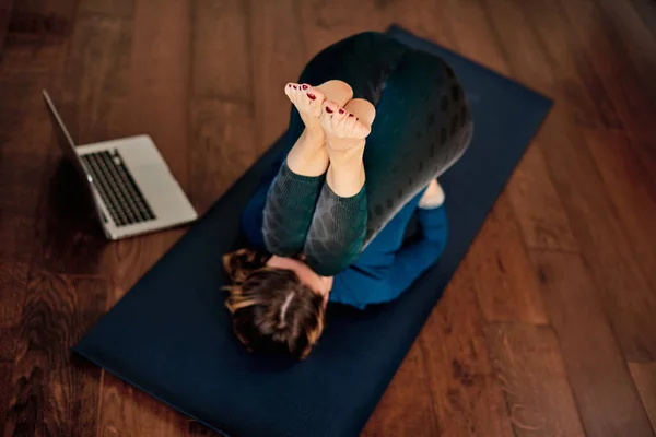 Aerial view of middle-aged woman lying down on a floor at home, doing yoga and following online class over laptop during lock down.