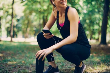 Sporcu bir kadın doğada çömelmiş, ara veriyor ve telefon kullanıyor..