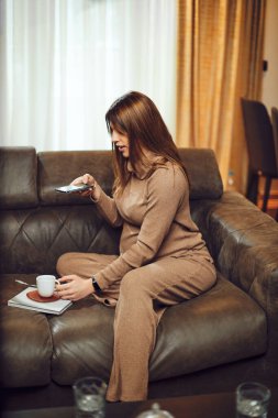 Hamile esmer kadın evdeki kanepede oturup telefonuyla sabah kahvesinin fotoğrafını çekiyor..