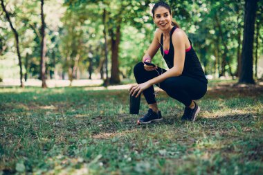 Sporcu kadın doğada çömeliyor ve koştuktan sonra mola veriyor. Sporcu kadın akıllı telefon kullanıyor ve içecek tutuyor.
