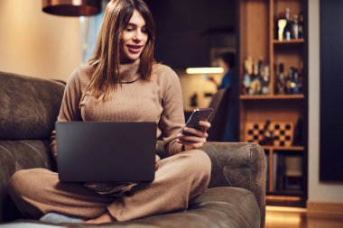 Hamile bir esmer kucağında dizüstü bilgisayarla kanepede oturuyor ve elinde telefonla internetten bir şeyler sipariş ediyor..