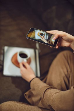 Hamile esmer kadın evdeki kanepede oturup telefonuyla sabah kahvesinin fotoğrafını çekiyor..