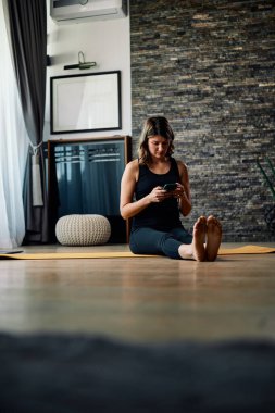 Orta yaşlı bir kadın yoga minderinde oturuyor ve telefonu internette sörf yapmak için kullanıyor..