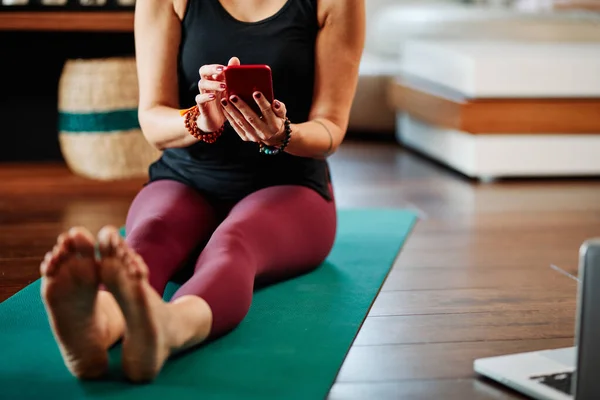 A woman sitting on the floor at home, using her phone and resting from yoga exercises.