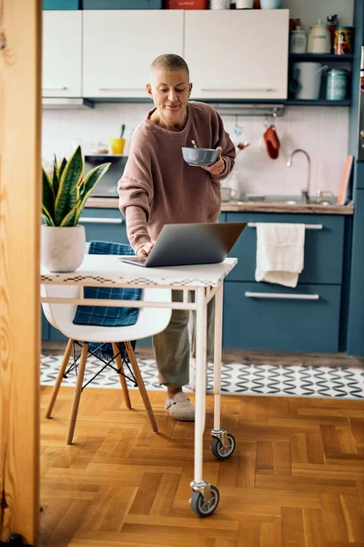 Smiling attractive mature woman standing in the kitchen at home, holding a bowl with cereal and typing something on the laptop. Morning routine.