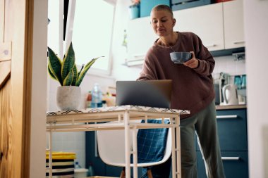 Gülümseyen olgun bir kadın mutfakta elinde mısır gevreği kasesiyle dizüstü bilgisayarına bir şeyler yazıyor. Sabah rutini.