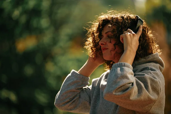 若いです巻き毛の女性が屋外に立って ヘッドフォンを置く — ストック写真
