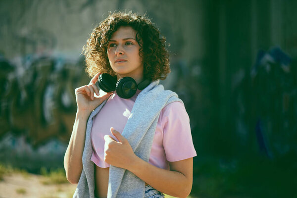 A young woman with curly hair standing outdoors and putting headphones on.