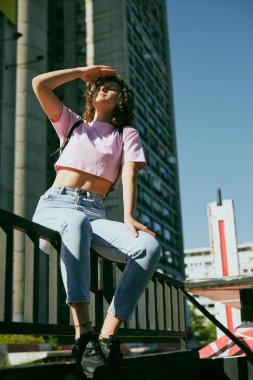 A pretty girl with curly hair sitting in urban part of the city, listening music and enjoying sunny day.