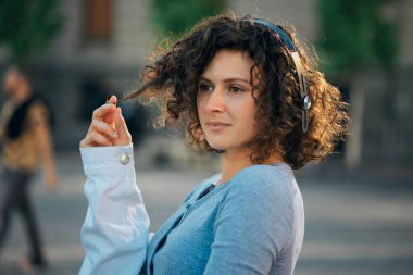 A young woman with curly hair standing on the street and listening music.