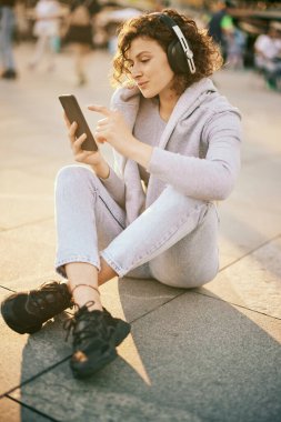Genç, mutlu bir kız, kulaklıkla kaldırımda oturuyor ve telefonda müzik seçiyor..